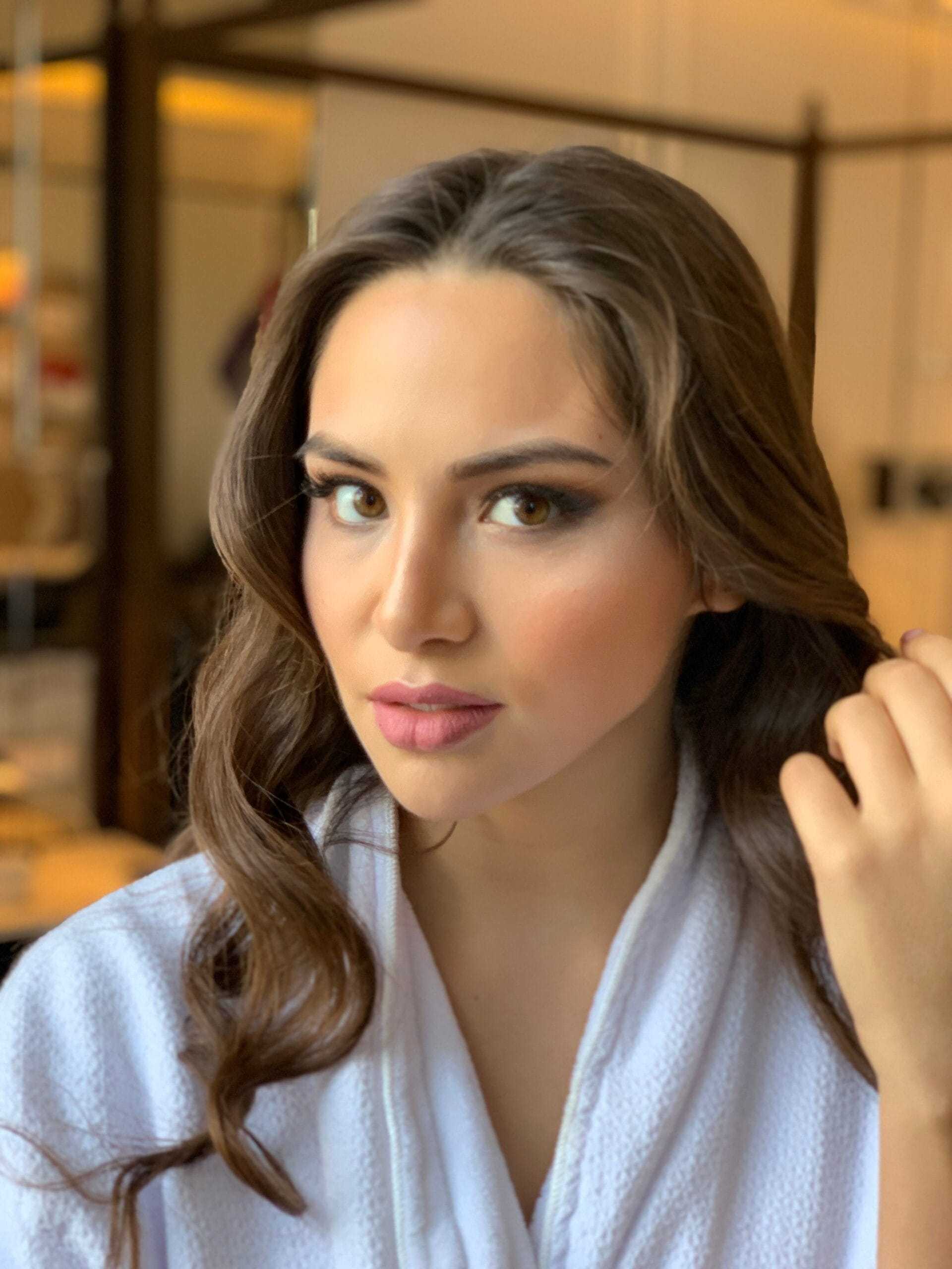 Young woman with long brown hair and makeup, wearing a white robe indoors.