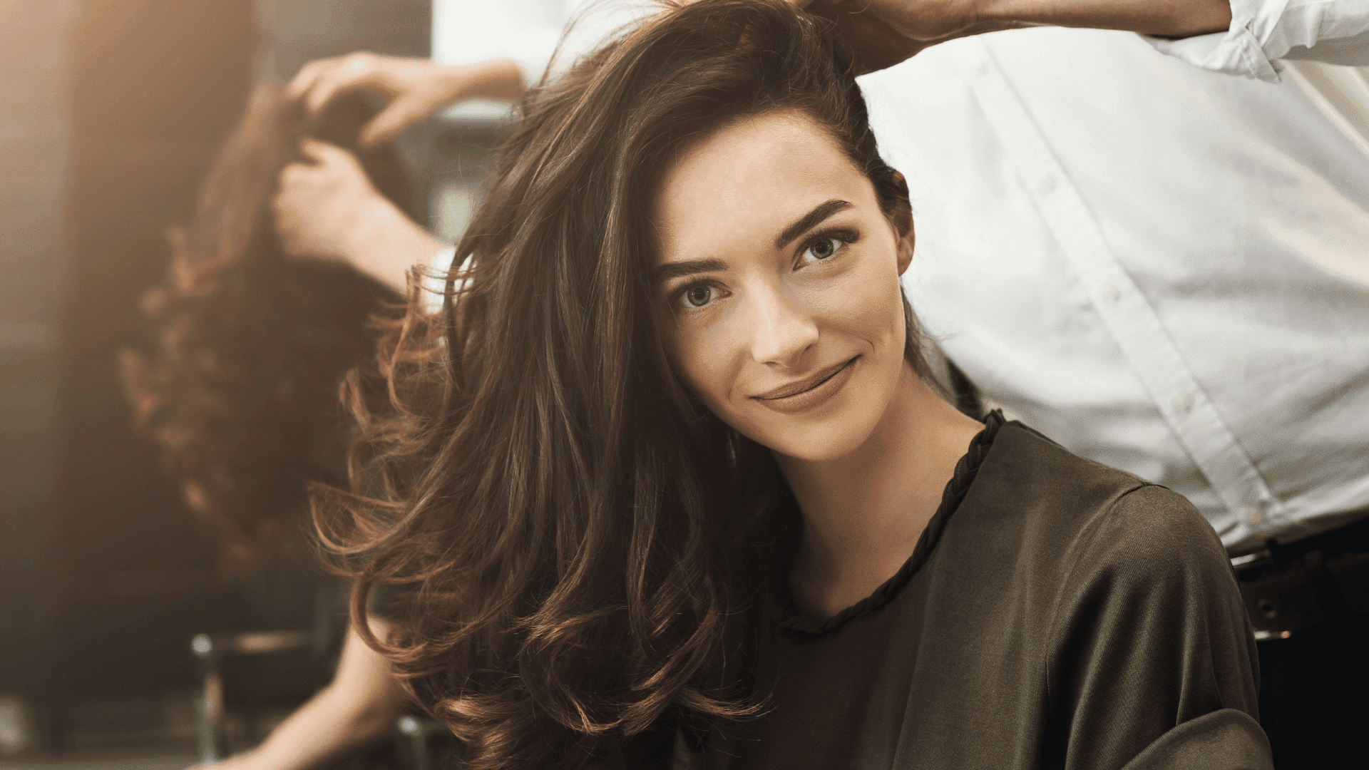 Hair stylist works on woman's long, wavy hair in a salon setting.