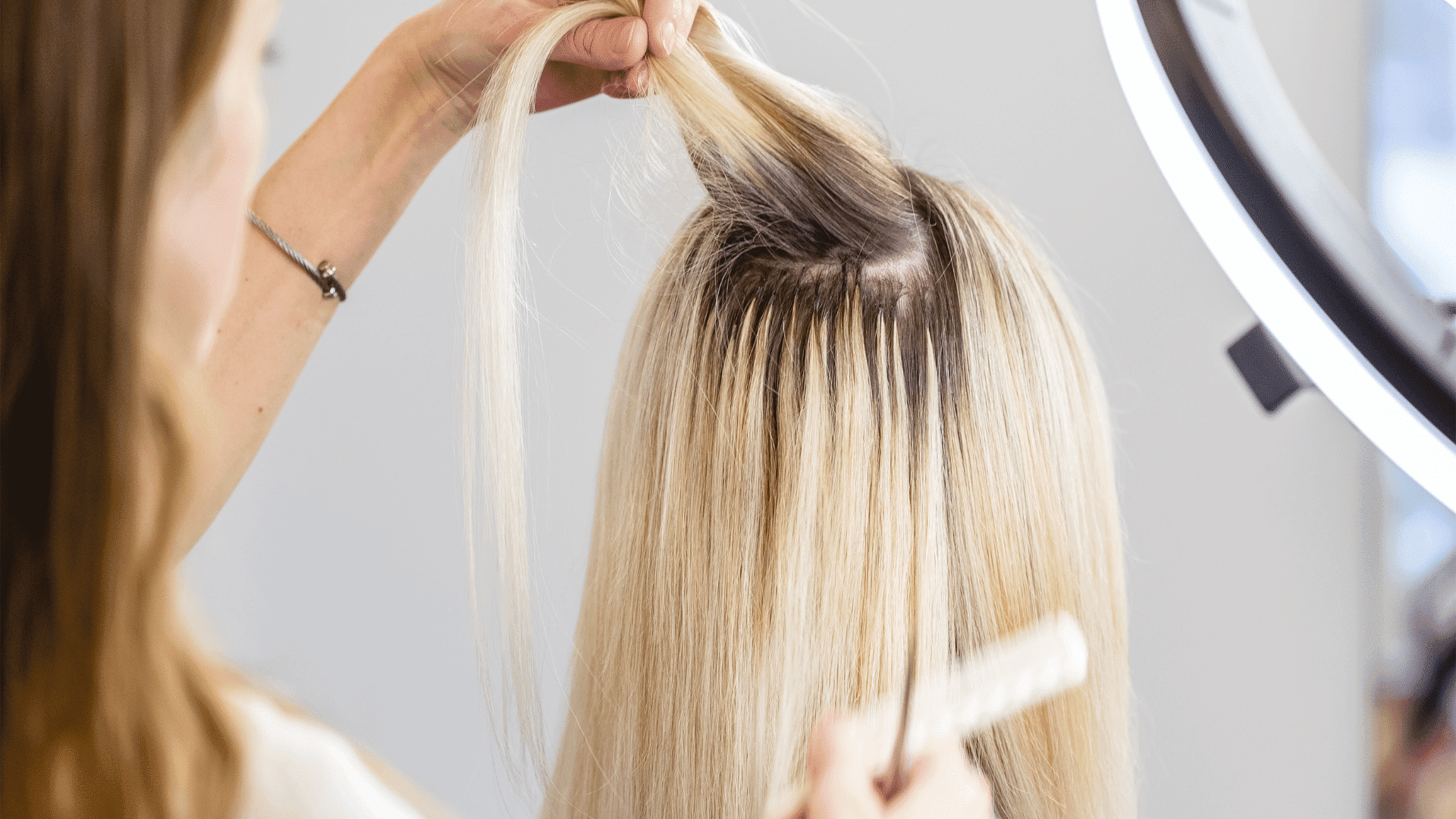Hair stylist applying extensions to woman's blonde hair under a ring light.