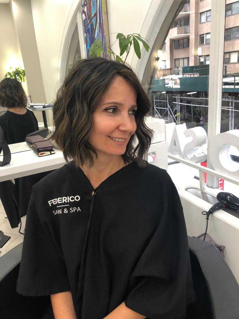 Woman with short wavy hair at a salon in front of large windows with a city view.
