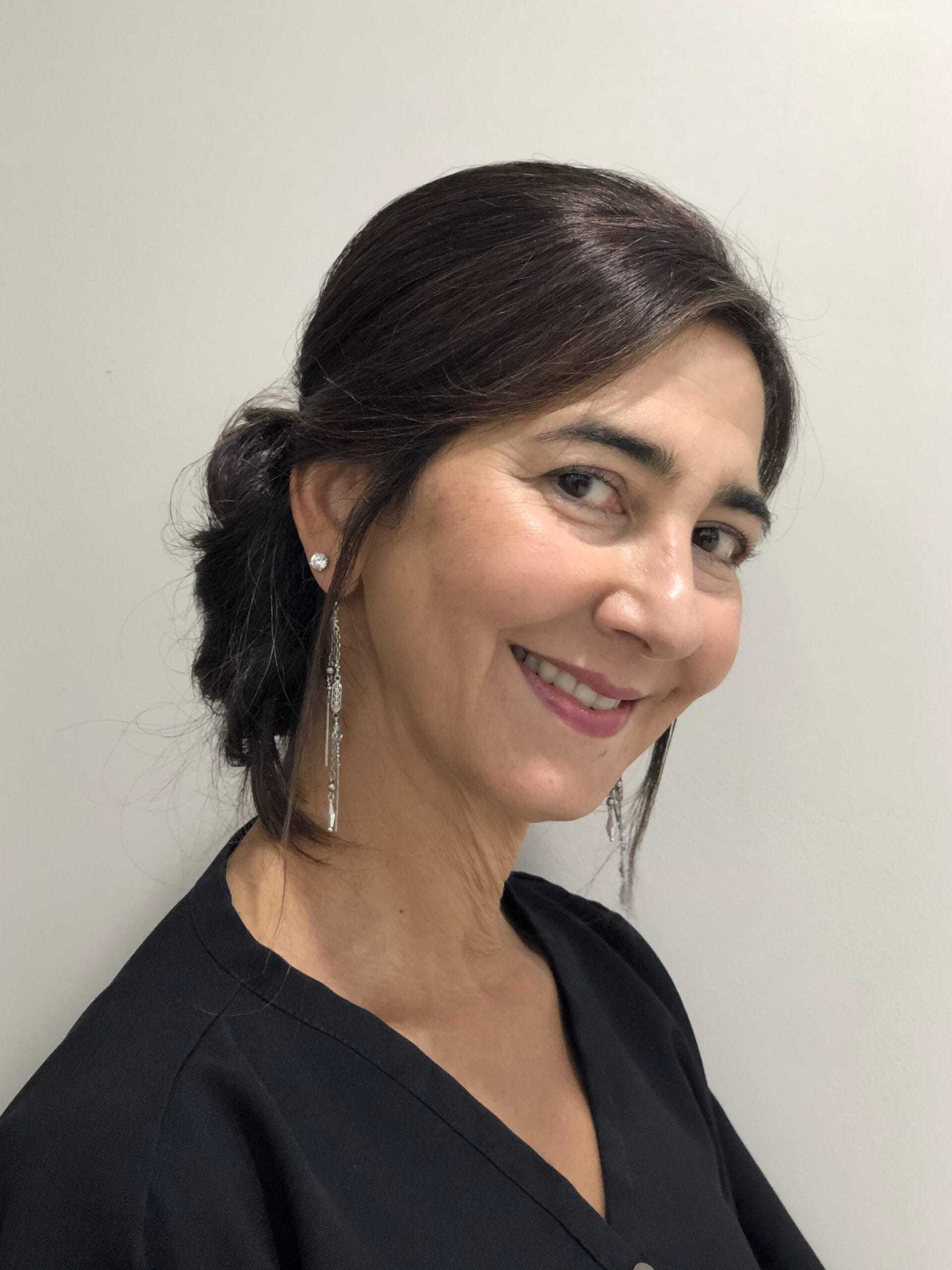 Woman with dark hair in a bun, smiling, wearing a black top and long dangling earrings.