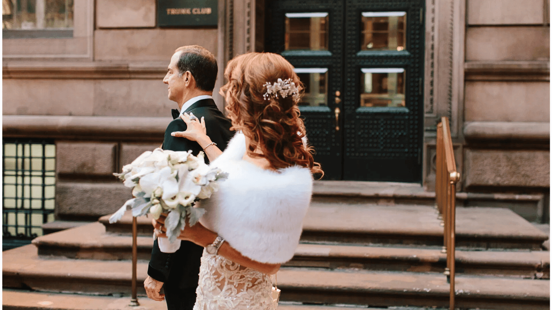 Bride in elegant gown and shawl approaches a man outside a building's entrance.