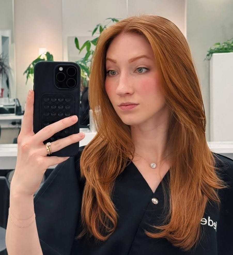 Young woman with long red hair takes a selfie in a salon, wearing a black gown.
