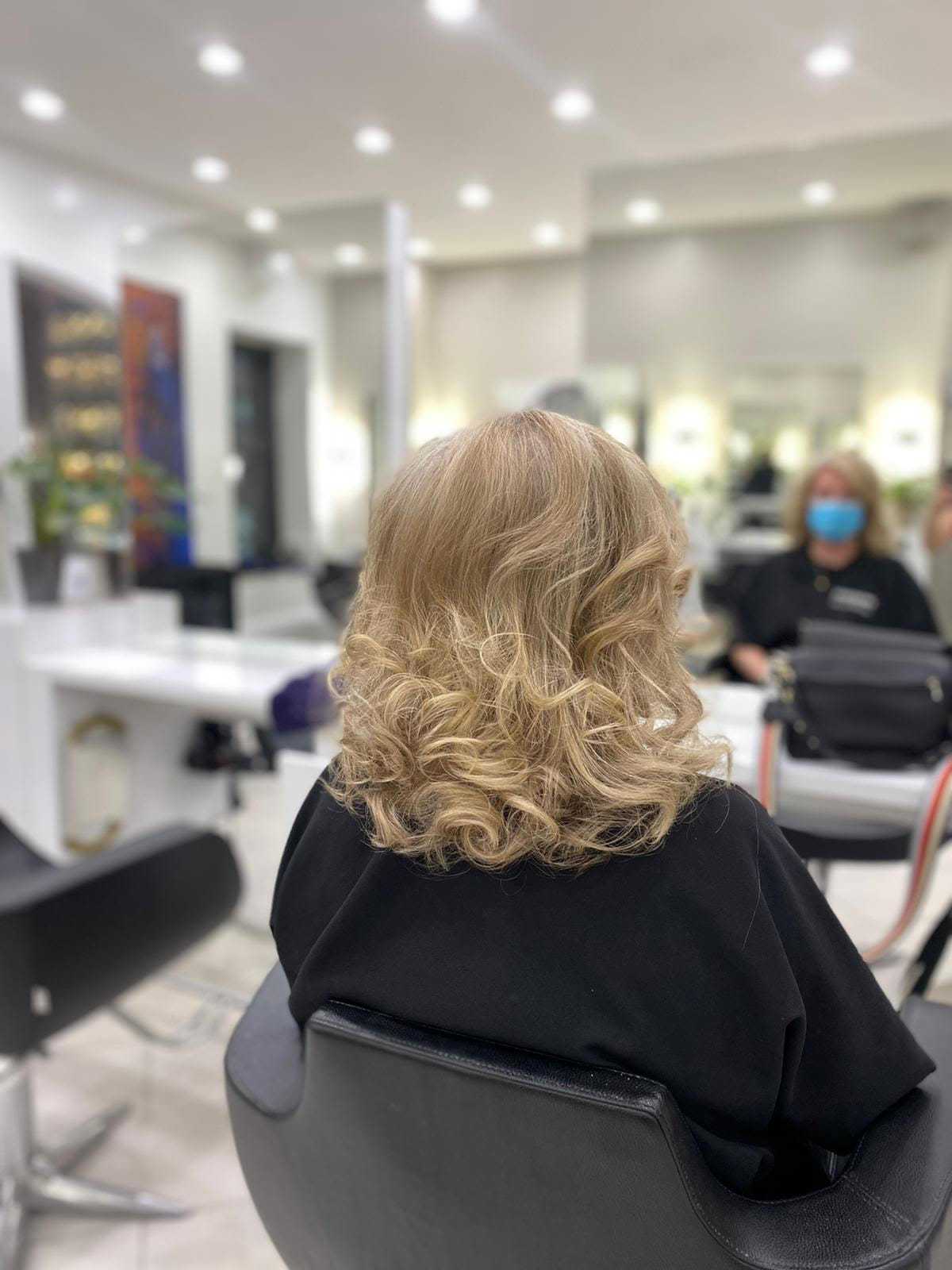 Woman with curly blonde hair sitting in a salon chair, mirrors and lights in the background.